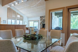 A modern dining area with a glass table surrounded by woven chairs, a stone fireplace, and large windows.