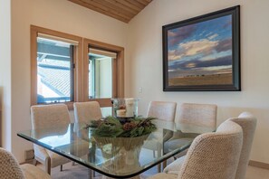 A dining area with a glass table surrounded by wicker chairs, a framed landscape painting on the wall, and a view through a window. - Wildflower Condo 617 - Spectacular 3rd Floor Penthouse with Pool Access (Sun Valley)
