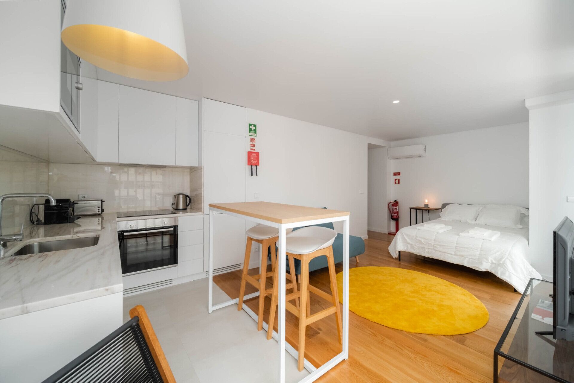 Una habitación de hotel moderna y compacta con kitchenette, una cama, un pequeño comedor y una alfombra amarilla.