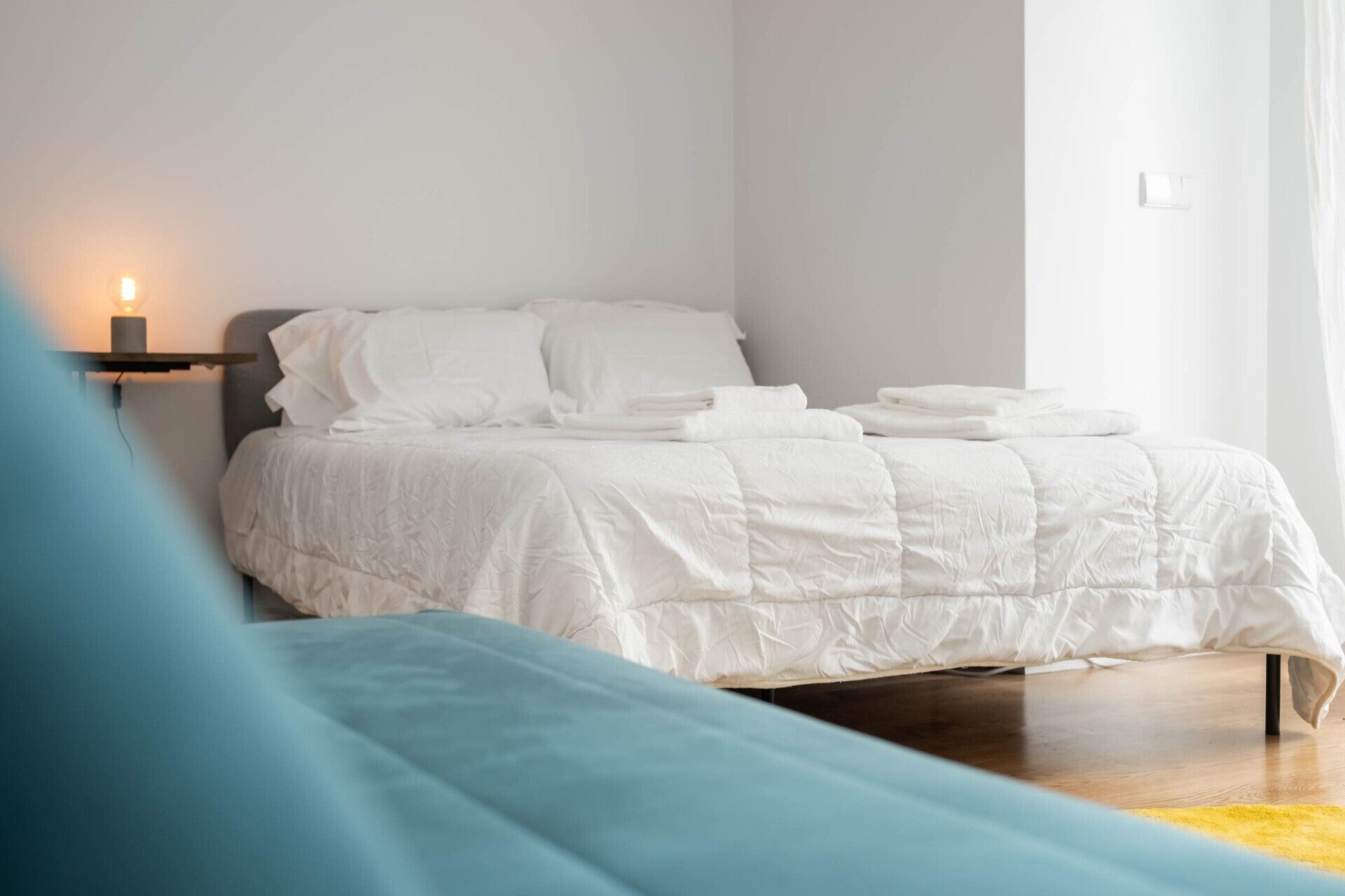 A neatly made bed with white bedding, a bedside table with a lamp, and a window with curtains.