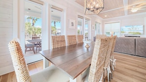 A spacious dining area with a wooden table and wicker chairs, large windows offering views of palm trees, and a ceiling fan. - Cottages of Romar 13 | 50 Steps to Beach Boardwalk! Amazing Home! Pool! (Orange Beach)