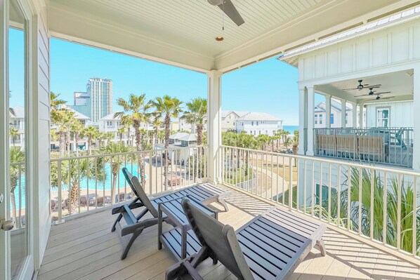 A covered balcony with two lounge chairs, a pool, and a view of nearby buildings and palm trees.