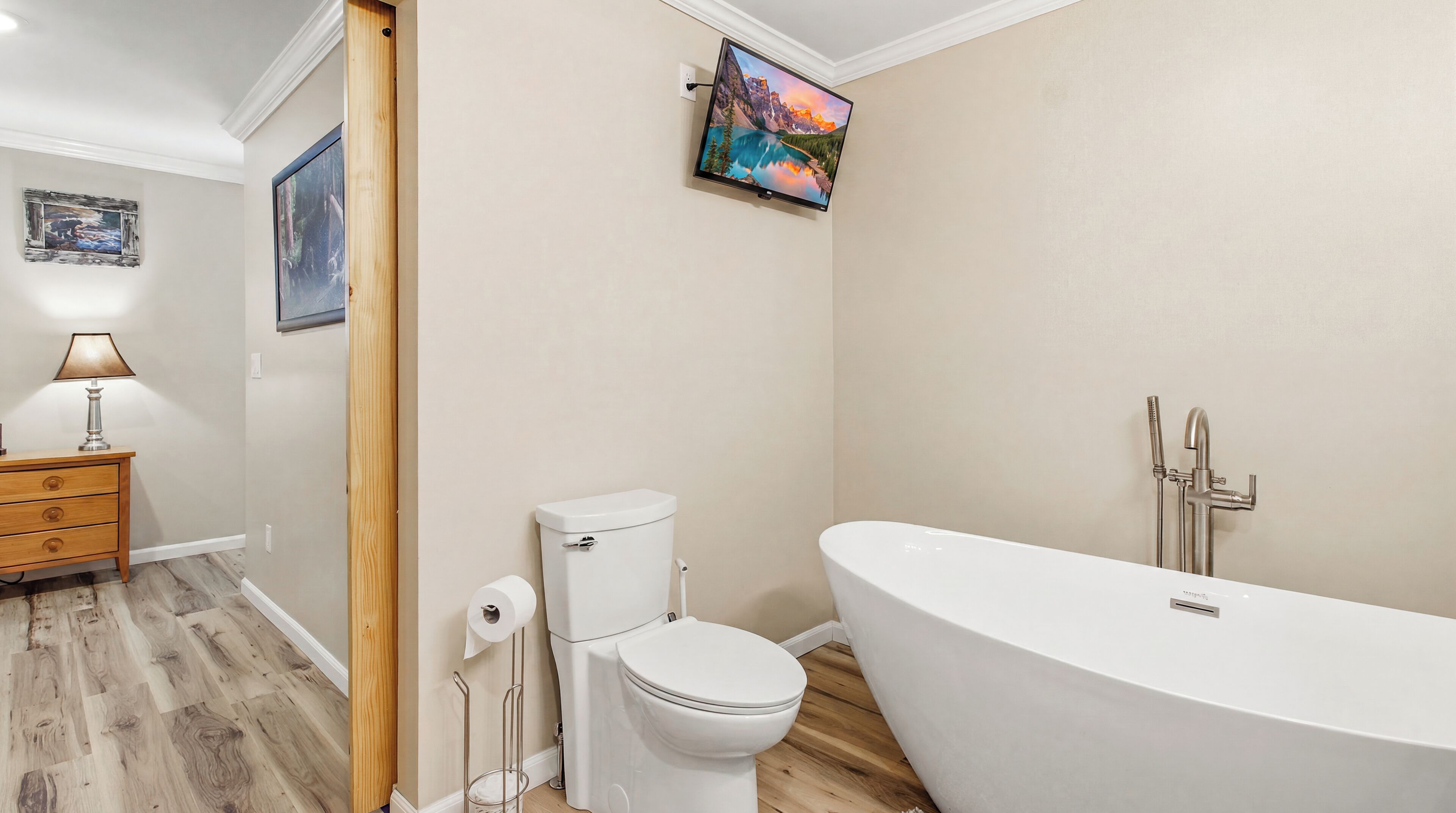Ein Badezimmer mit Toilette, einer Badewanne, Holzboden, einem am Wand montierten Fernseher und einem Nachttisch mit Lampe.