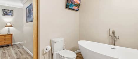 A bathroom with a toilet, a bathtub, a wooden floor, a wall-mounted TV, and a wooden nightstand with a lamp.