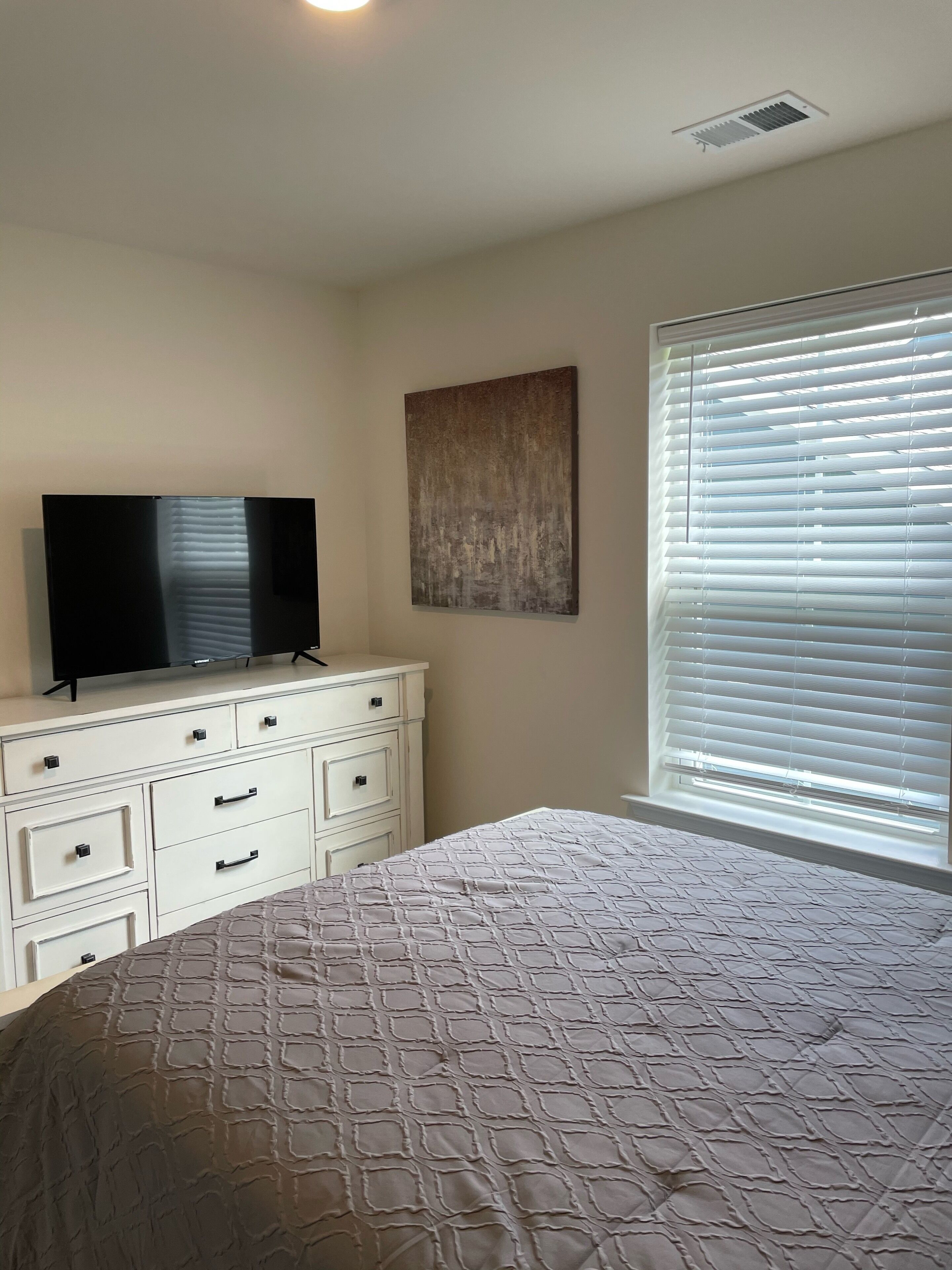 Un dormitorio con cama, cómoda, televisor de pantalla plana, un cuadro y una ventana con persianas.