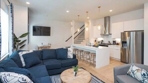 Una sala de estar moderna con un sofá modular, una mesa de centro redonda y un sillón gris. Se ve una cocina al fondo con electrodomésticos de acero inoxidable y una barra.