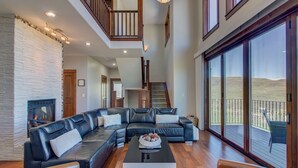 A spacious living area with a large sectional sofa, a coffee table, and a fireplace. There is a staircase leading to an upper level with large windows offering a view of the outdoors.