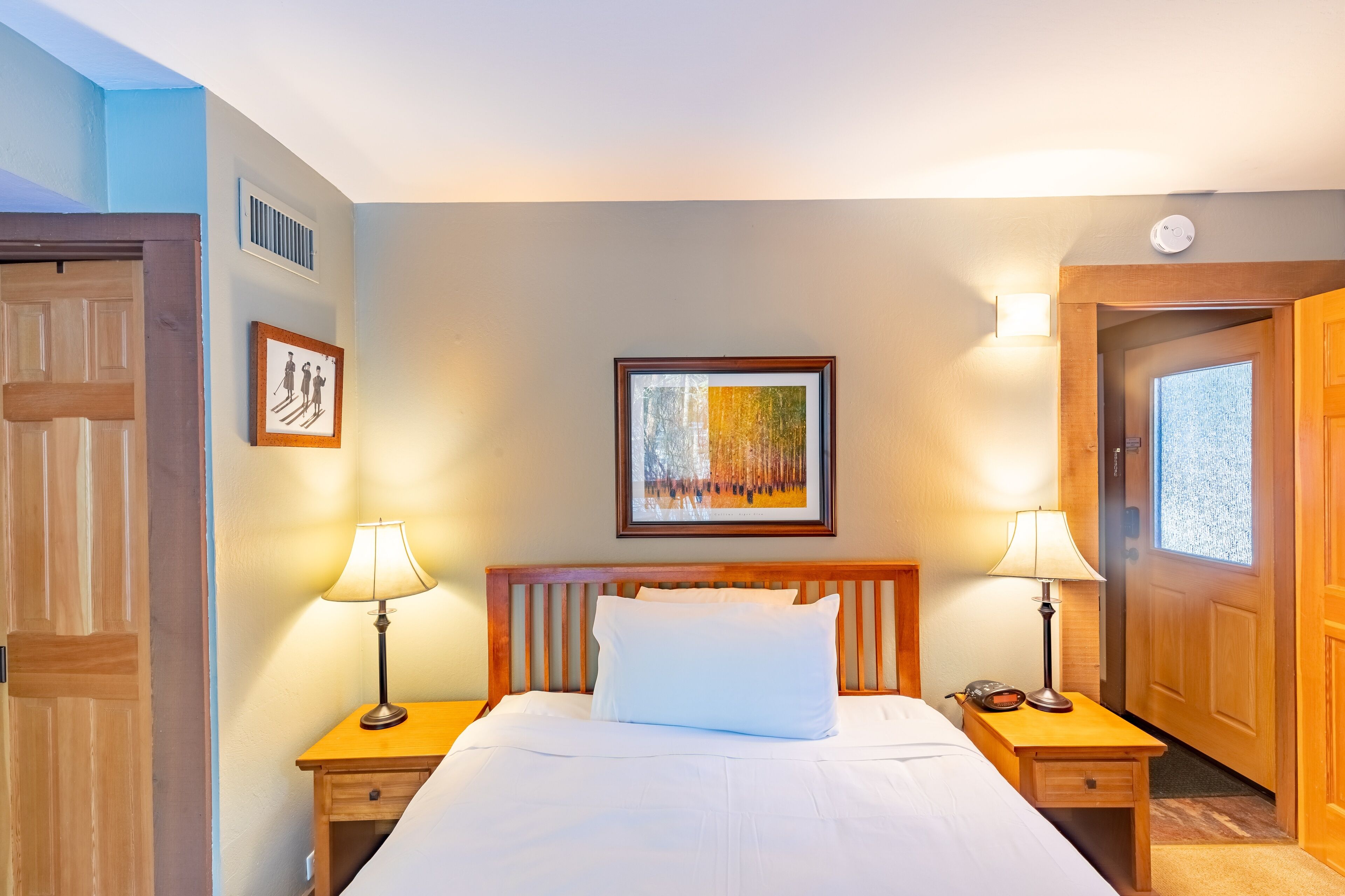 A hotel room with a large bed, two bedside tables with lamps, a framed picture above the bed, and a door to the right.