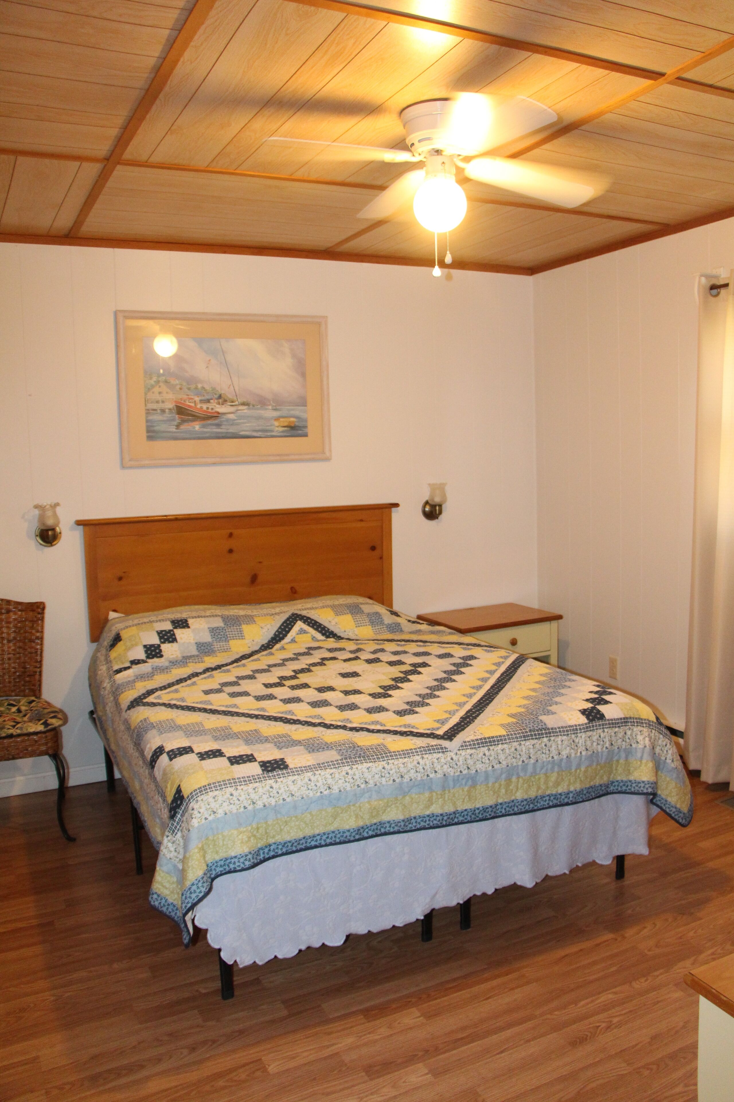 A bedroom with a wooden ceiling, a bed with a quilt, a nightstand, a chair, and a painting on the wall.