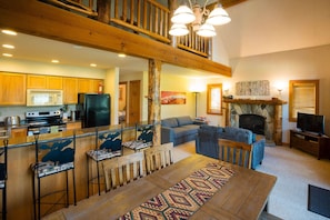 A spacious living area with a high ceiling, a fireplace, a television, and a dining area with a patterned rug.