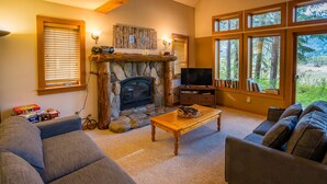 A cozy living room with a stone fireplace, a television, and a large window with a view of trees. - Crazy Squirrels Cabin at Timberline Meadows (Winthrop)