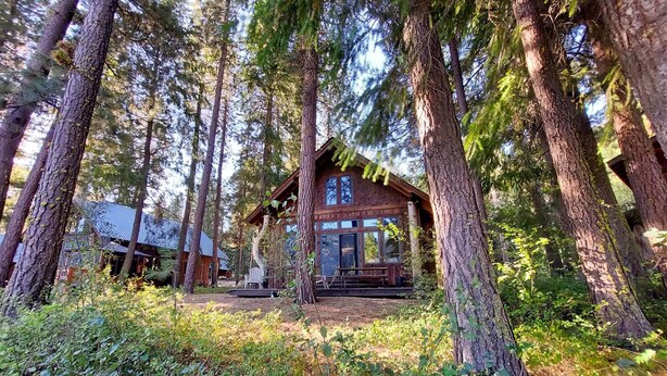 A cabin surrounded by tall trees, with a porch and a large window. - Crazy Squirrels Cabin at Timberline Meadows (Winthrop)