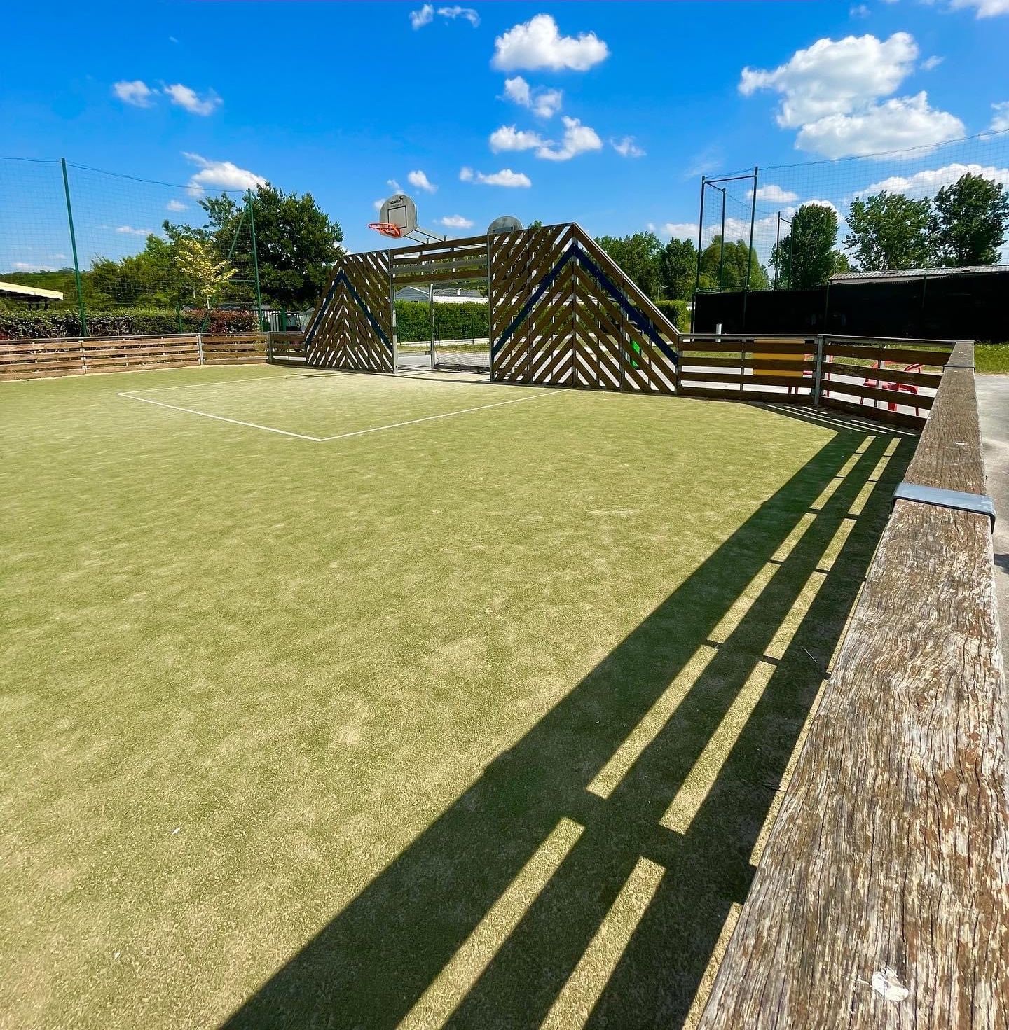 Ein Sportplatz im Freien mit einem Basketballkorb, umgeben von einem Holzzaun und einer überdachten Sitzgelegenheit.