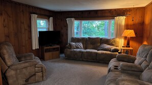 A cozy living room with a large sectional sofa, two armchairs, a flat-screen TV on a wooden stand, and a lamp on a side table.