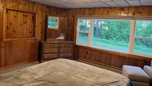 A bedroom with wooden walls, a large bed, a dresser, and a window with a view of trees.