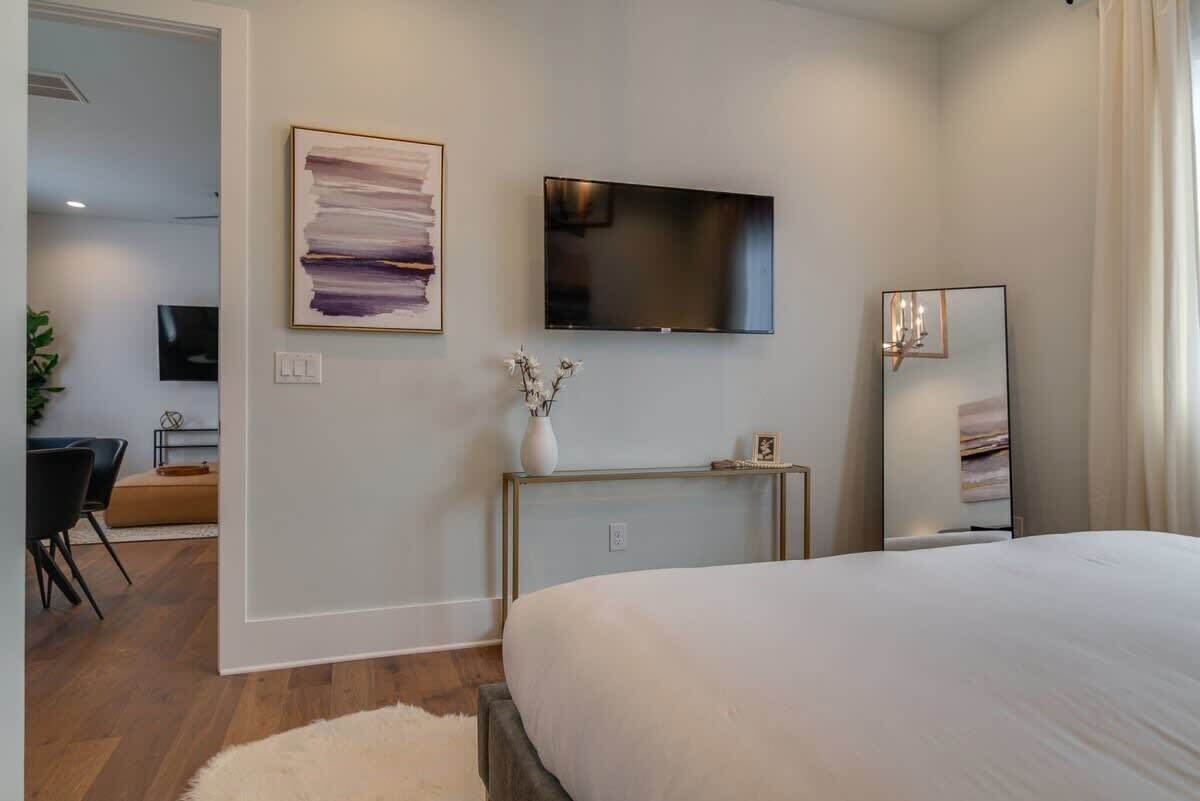 Een moderne slaapkamer met een groot bed, een flatscreen televisie aan de muur, een nachtkastje met een vaas en een spiegel.