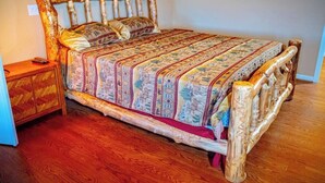 A wooden bed with a patterned bedspread and pillows, a nightstand with a drawer, and a wooden floor. - Royal Ranch (Cañon City)