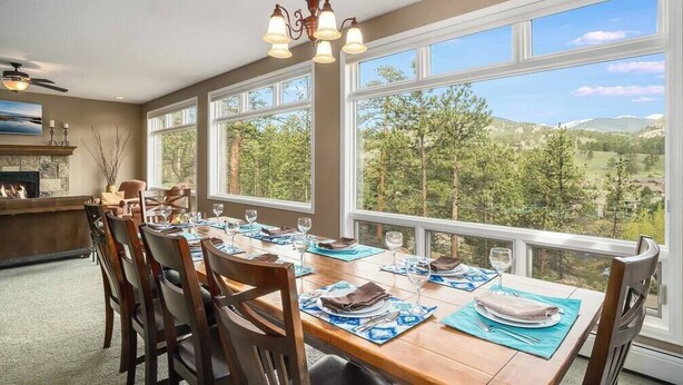 Comedor con una gran mesa dispuesta para seis comensales, ventanas amplias con vista a un paisaje boscoso y una chimenea de piedra.