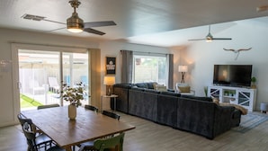A living room with a wooden dining table, green chairs, a grey sofa, and a TV on a white cabinet. - Prickly Pear Desert Oasis- 3 Bedroom/Pool/Spa/Putting Green (Glendale)
