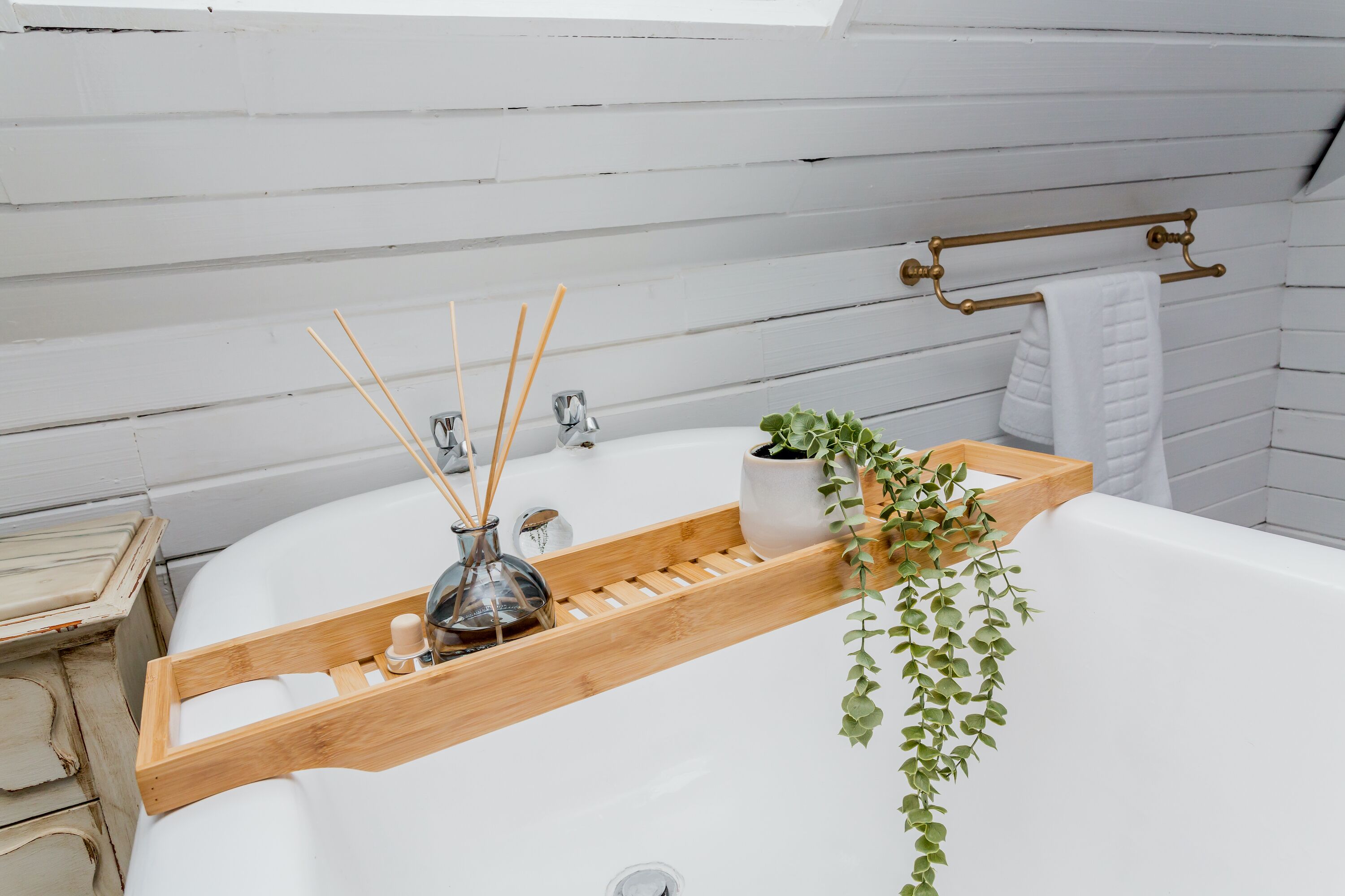 Ein Badezimmer mit einer weißen Badewanne, einem Holztablett mit einer Glasflasche und Schilff Diffusoren, einer Topfpflanze und einem Handtuchhalter.