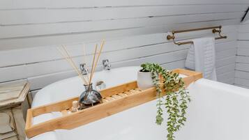 Ein Badezimmer mit einer weißen Badewanne, einem Holztablett mit einer Glasflasche und Schilff Diffusoren, einer Topfpflanze und einem Handtuchhalter.