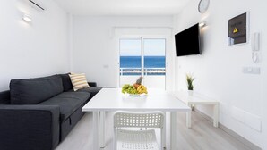 A modern, minimalist room with a white sofa, a dining table with a fruit bowl, a white chair, a TV mounted on the wall, and a view of the sea through a large window.