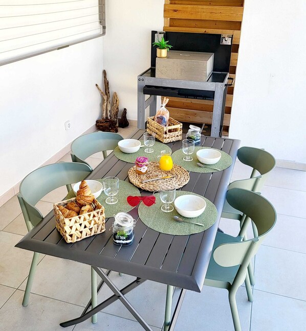 A dining table set with plates, glasses, and a basket of croissants, surrounded by green chairs. - Somptueux Appart Neuf 3 Pièces en rdc Tout Confort Avec Accès Plage et Piscine (Linguizzetta)