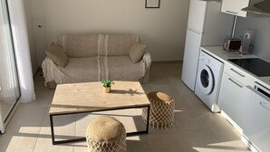 A modern living room with a sofa, a wooden table, a washing machine, and a kitchen area.