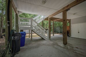 A covered outdoor area with a wooden staircase leading to a deck, a garage door, and a trash bin.