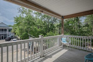 A covered porch with a white railing, a wicker chair with a cushion, and a view of a residential area with trees and a parked car.