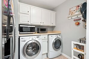 Une cuisine compacte avec des meubles blancs, un four à micro-ondes, un lave-linge et une étagère ornée d’objets décoratifs.