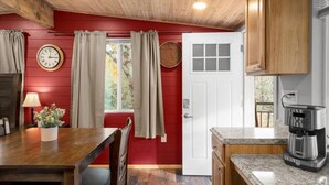A kitchen with a red wall, wooden ceiling, a white door, a clock, a coffee maker, and a dining table with chairs. - 19 Creekside Retreat (Sedona)