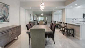 A modern dining area with a long table, chairs, and a kitchen in the background.