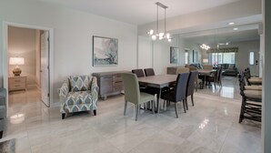 A modern dining area with a large table, chairs, and a chandelier. There is a sofa and a lamp in the background.