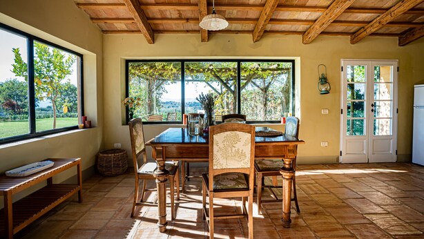 A dining area with a wooden table and chairs, large windows with a view of greenery, and a rustic ceiling. - Villa il Sogno - Homelike Villas (Ostra Vetere)