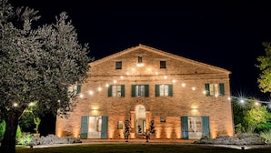 A large, illuminated brick building with a symmetrical facade, multiple windows, and a central entrance. - Villa il Sogno - Homelike Villas (Ostra Vetere)