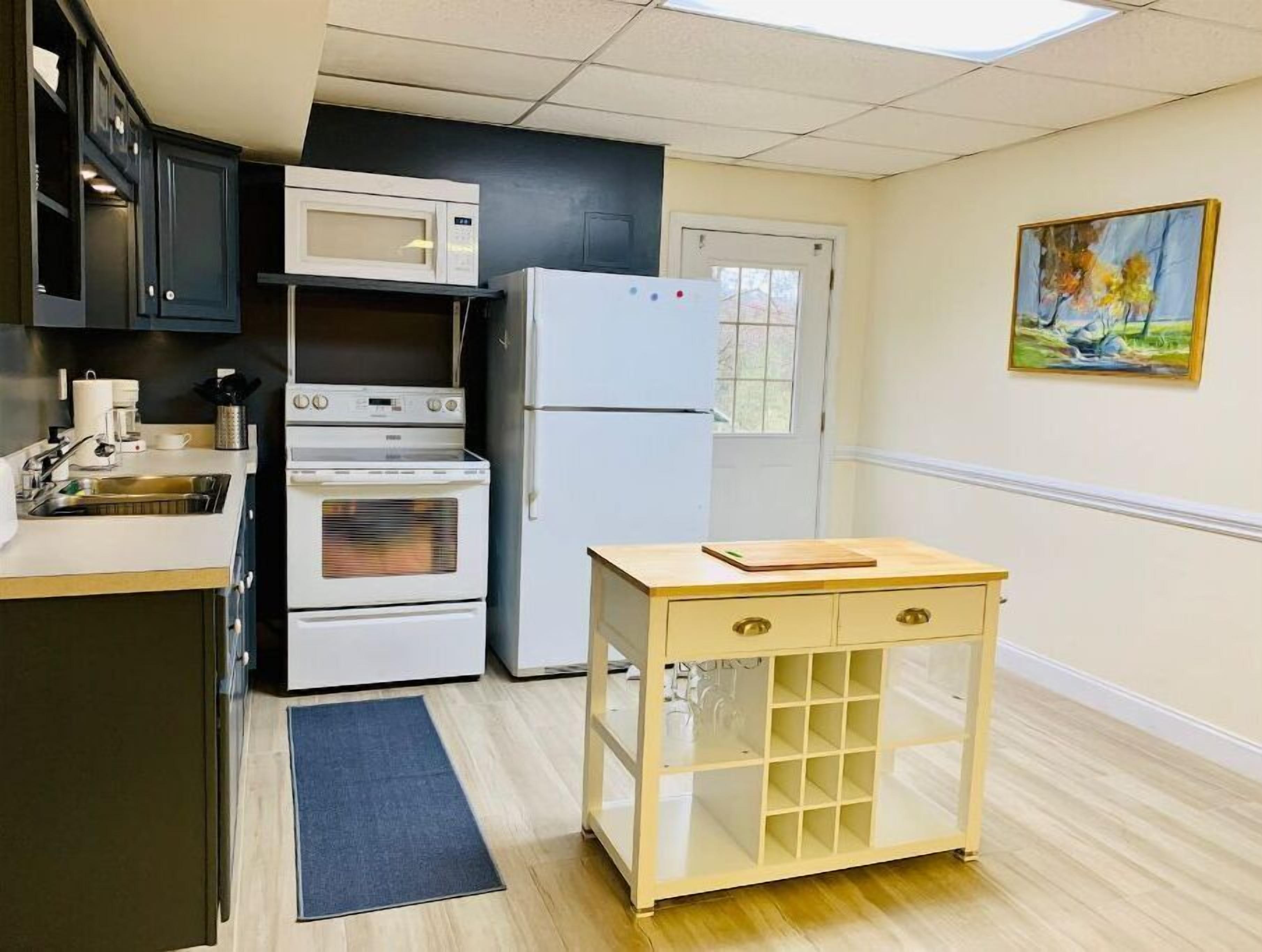 A compact kitchen with a microwave, oven, and refrigerator, a small wooden table, and a painting on the wall.