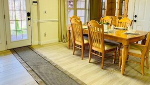 A dining room with a wooden table and chairs, a window with curtains, and a door with a doormat.