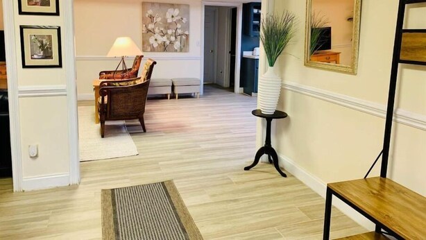 A hallway with a wooden floor, a wicker chair, a lamp, a mirror, and framed artwork on the walls.
