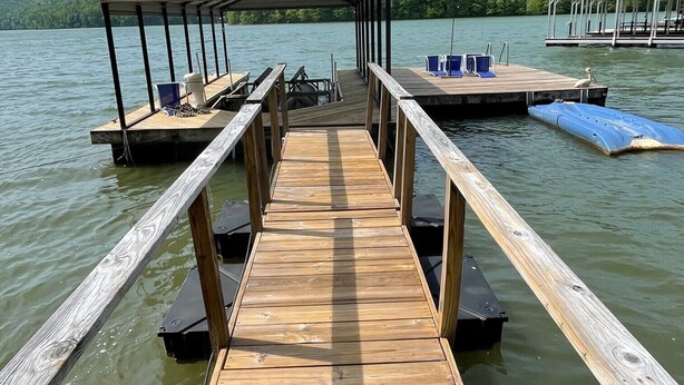 Un muelle de madera que lleva a un espacio de descanso cubierto con mesas y sillas, con vista a un lago rodeado de vegetación.