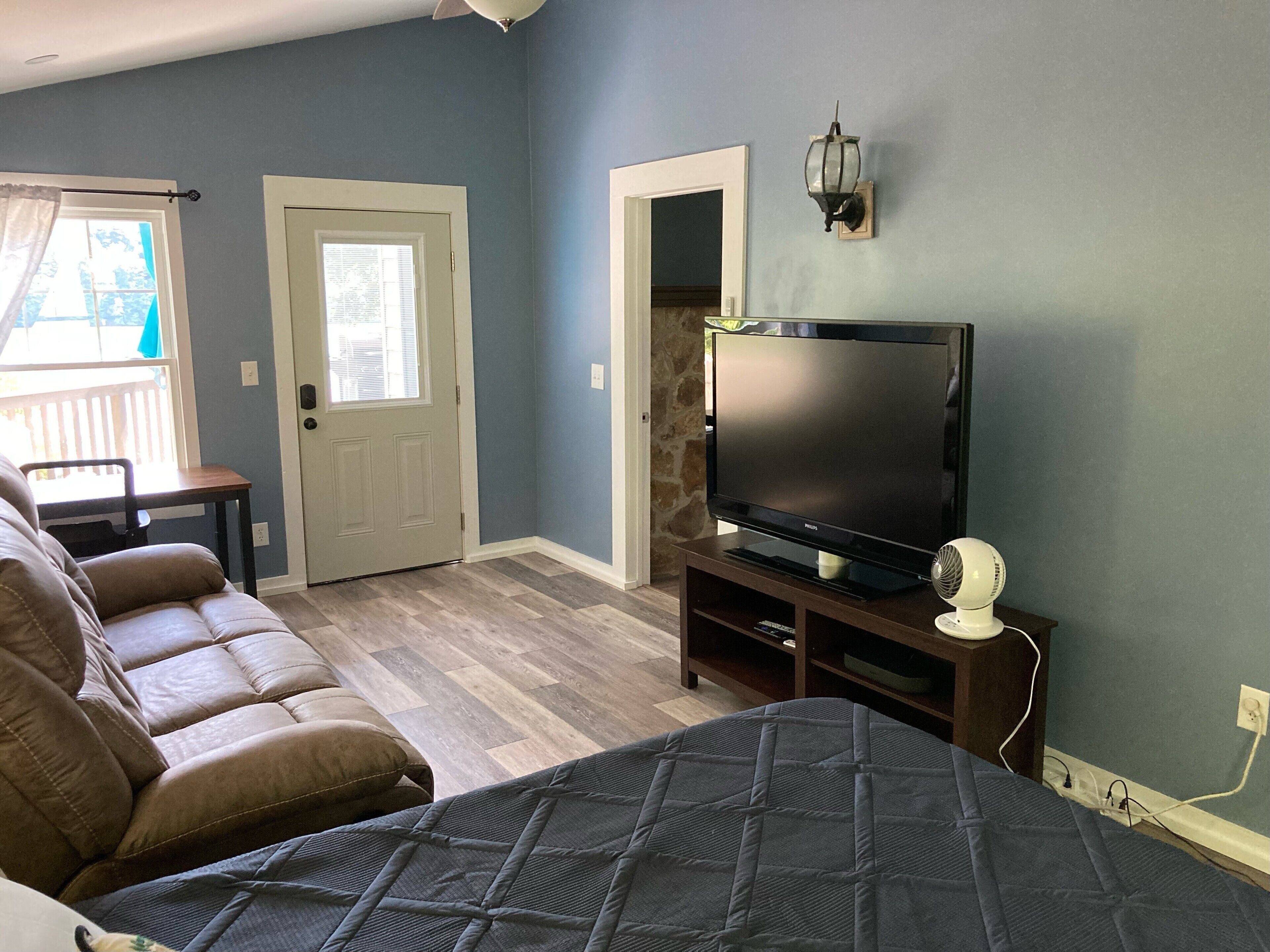 A living room with a large flat-screen TV on a wooden stand, a brown leather sofa, and a blue quilted ottoman.