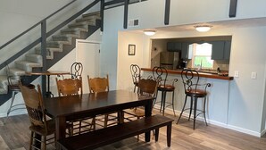 Un comedor con mesa y sillas de madera, una escalera y una zona de cocina con refrigerador y pileta.