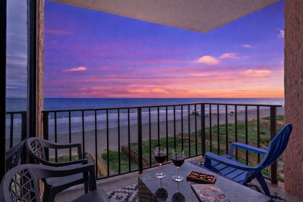 Un balcon avec vue sur la plage et le coucher de soleil. On y trouve deux chaises et une table avec deux verres de vin rouge.