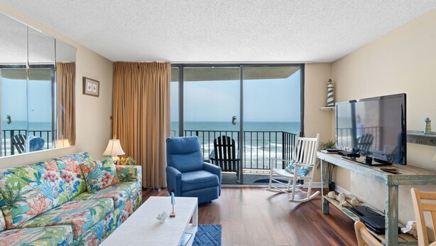 Ein Zimmer mit einer Couch, einem blauen Sessel, einem Schaukelstuhl, einem Fernseher und Blick auf den Ozean.
