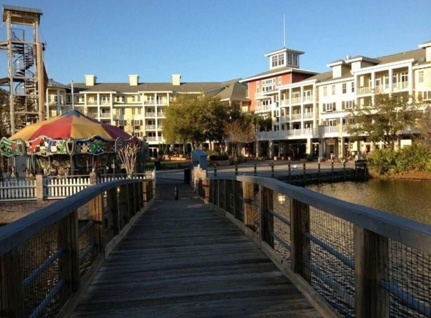 A wooden boardwalk leading to a waterfront area with a carousel and multi-story buildings in the background.