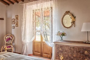 Une pièce avec une porte en bois, une chaise recouverte d’un tissu à motifs floraux, une commode à tiroirs, un miroir avec un cadre orné de motifs floraux et un vase rempli de fleurs.