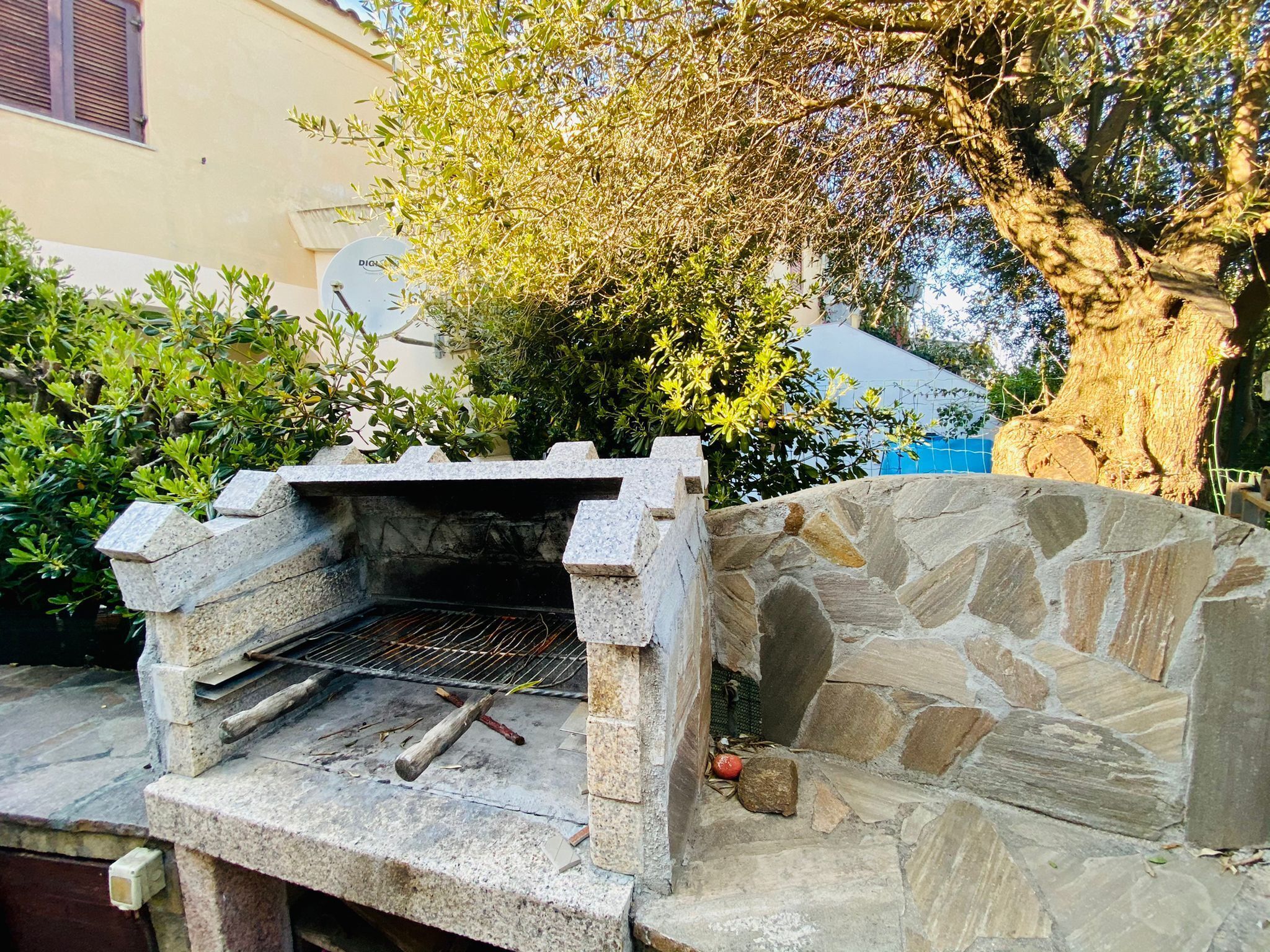 An outdoor stone-built grill area with a metal grate, surrounded by stone walls and greenery.