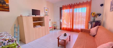 A living room with a sofa, a television on a stand, a table with a vase of flowers, and a window with curtains.
