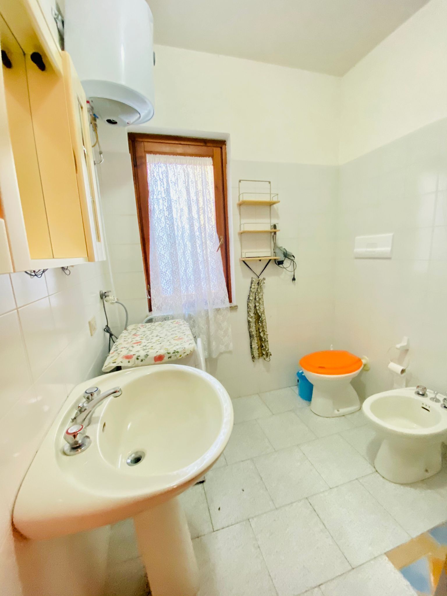 A bathroom with a sink, toilet, bidet, and a window with lace curtains.