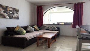 Una sala de estar con sofá, cojines, una mesa de madera y un ventanal con cortinas.